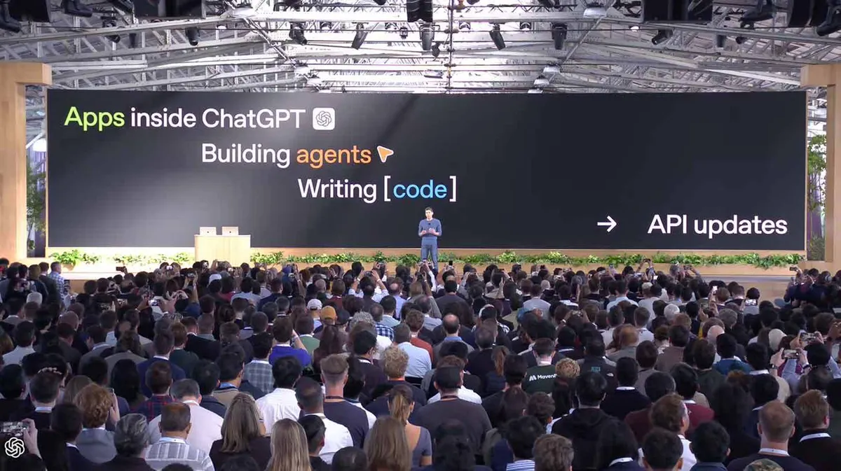Saalitäis inimesi kuulamas esitluskõnet. Laval seisab mees ja tema taga on suur ekraan, kus on kirjas: "Apps inside ChatGPT", "Building agents", "Writing [code]" ja "API updates". Lava ääres on palju taimi.