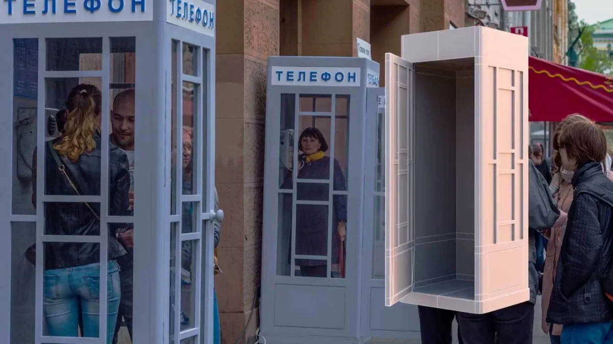Tänavavaade mitme telefoniputkaga, kus inimesed räägivad telefoniga. Üks telefoniputkadest on ilma seinte ja külgedeta, eksponeerides selle siseruumi. Taustal on näha punase varikatusega hooneid.