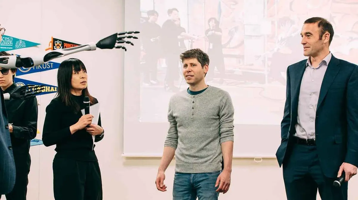 Kolm inimest on toas, naine räägib mikrofoni, käes paberid. Taustal näha projitseeritud pilti ja seinal lipukesi erinevate ülikoolide logodega. Robotkäsi ulatub vasakult kaadrisse.